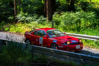 Red Toyota Celica GT-Four ST185 driving through Sturt Valley during the Adelaide Rally, with pop-up headlights raised and surrounded by forest vegetation and guard rails.