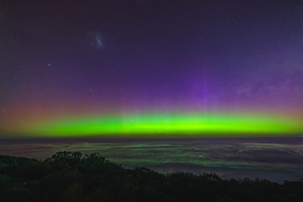 Southern Lights with green and purple aurora bands above the ocean at Muhan Viewpoint, with the Large Magellanic Cloud visible.