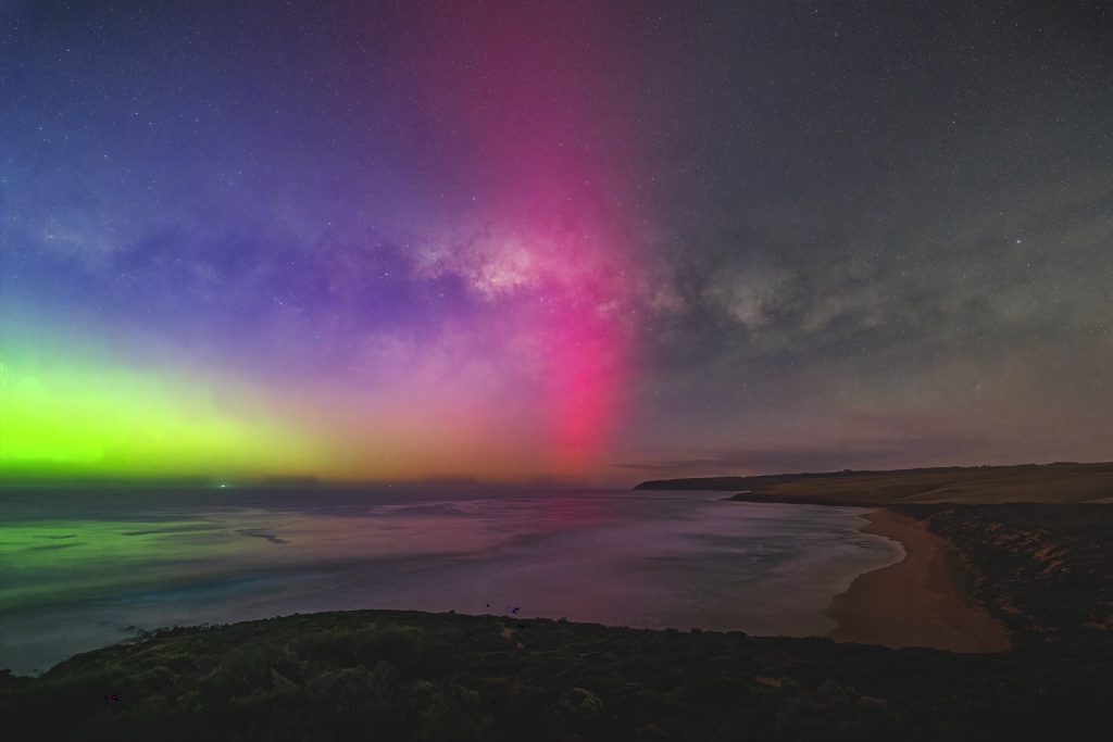Milky Way arch above Parsons Beach with a bright pink aurora beam and green aurora glow rising from the southern horizon.