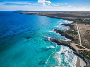Aerial view of Sleaford Bay in Lincoln National Park, showing turquoise coastal waters, rugged cliffs, white sand beach and winding dirt access road under clear blue skies.