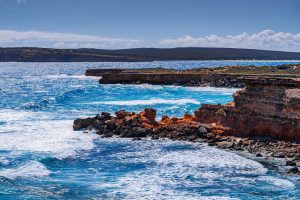 Rugged red sandstone cliffs and turquoise ocean waves along the coastline of Venus Bay on the Eyre Peninsula, with jagged rock formations and white sea foam under a bright blue sky.