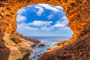 View from inside Woolshed Cave on the Eyre Peninsula, showing orange sandstone walls framing the blue ocean, tide pools and coastal rock formations under a partly cloudy sky.