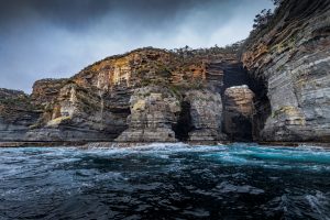 Dramatic layered sea cliffs with a natural rock arch along the Tasman Island coastline, photographed from the ocean under stormy skies in Tasmania.