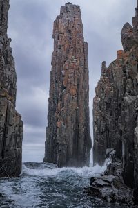 A tall vertical sea stack rising between narrow coastal cliffs at Tasman Island, with waves crashing below and cloudy skies overhead.