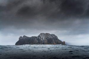 Tasman Island surrounded by rough ocean waves under dark storm clouds, photographed from sea level.