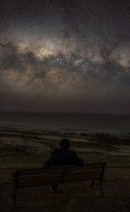 A person sitting beneath the bright Milky Way core at Haycock Point Lookout in Carrickalinga, South Australia, overlooking the coast at night.